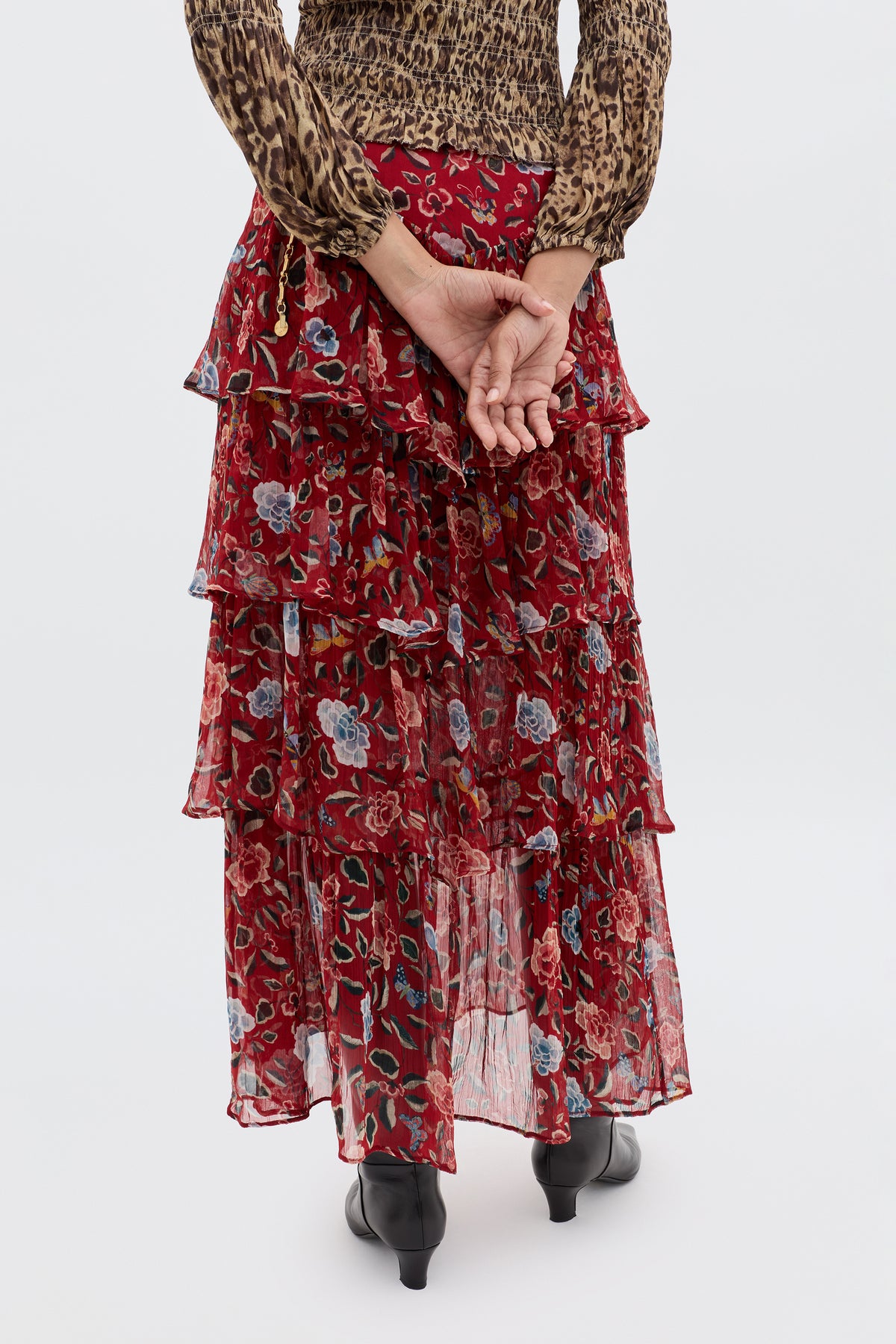 A person stands with hands clasped behind their back, wearing the Taryn - Butterfly Floral Burgundy skirt, black boots, and a long-sleeved animal print top, photographed from behind against a plain background.