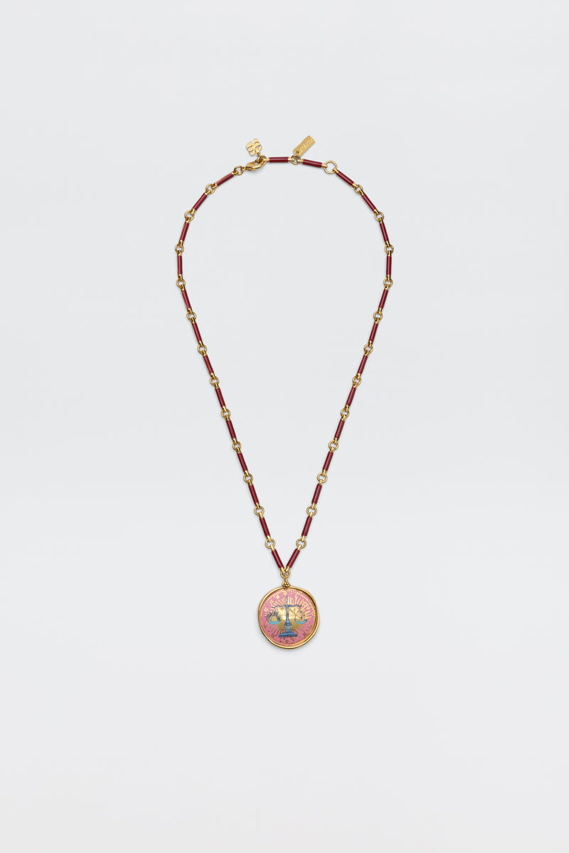Ari - Astro Libra gold necklace on a link chain featuring maroon beads and a round pendant with a scale icon against a pink sunburst background.