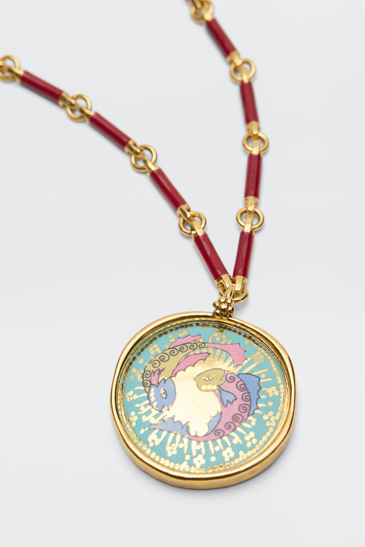 The Ari - Astro Pisces gold zodiac necklace features striking red links and a round pendant showcasing a colorful, stylized fish against a teal background with elegant gold accents. This eye-catching statement disc charm brings an artistic flair to any outfit.