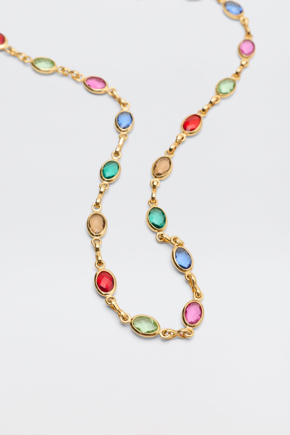 The Annabeth - Multi is a gold chain necklace adorned with evenly spaced oval-shaped gemstones in vibrant shades of red, blue, green, and pink, elegantly displayed on a light background.