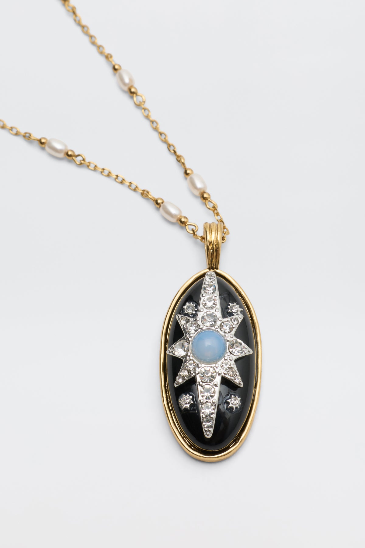 Andi - Black: A gold necklace with pearls and an oval pendant featuring a black background, a gold rim, and a star-shaped design adorned with white gemstones and a blue center stone.