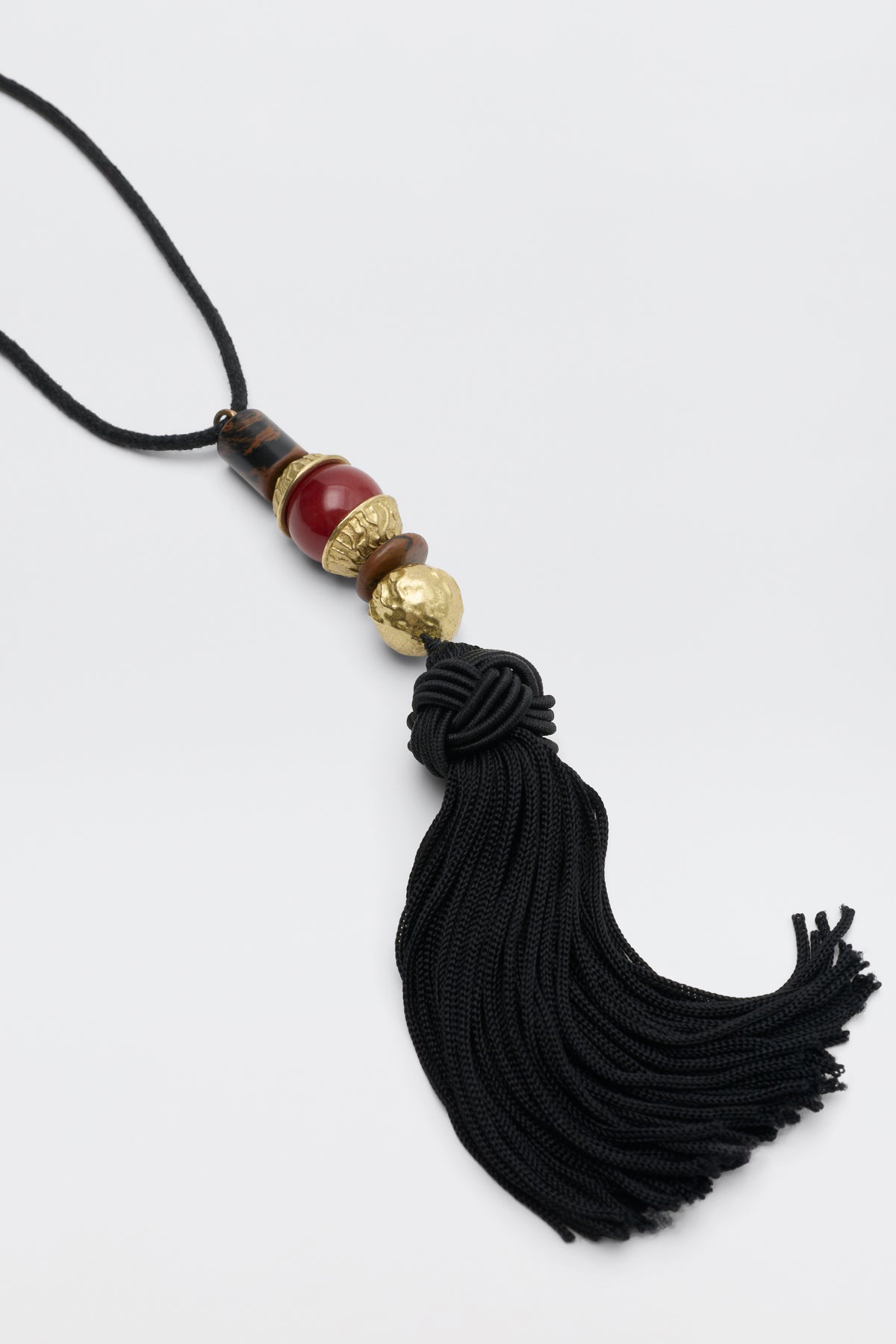The Rossa - Black necklace features a black cord, dark wood beads, red stone, hammered gold accents, and a black tassel.