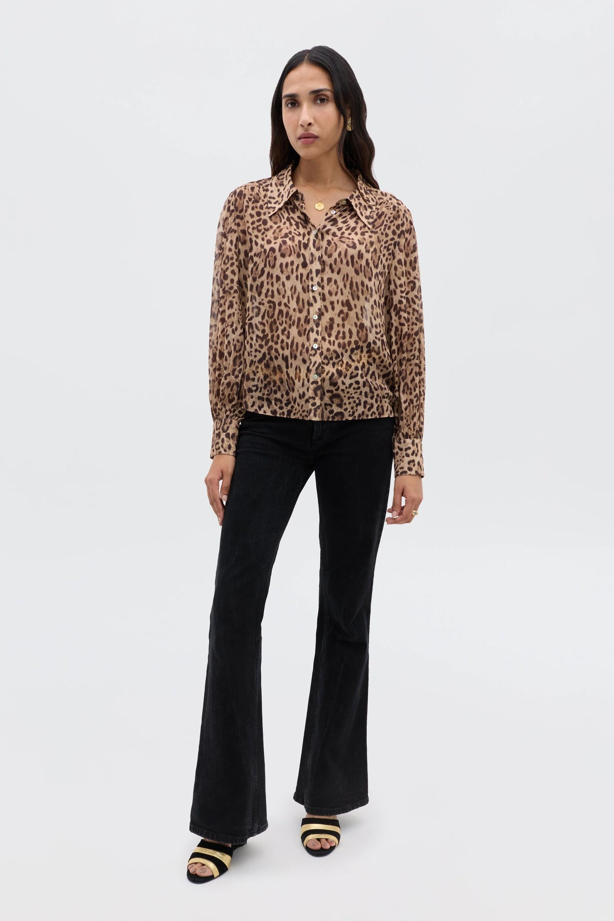 A woman with long dark hair and a neutral expression stands against a white background, wearing the Blake - Mila Leopard Brown top, black flared pants, and black open-toe heeled sandals.