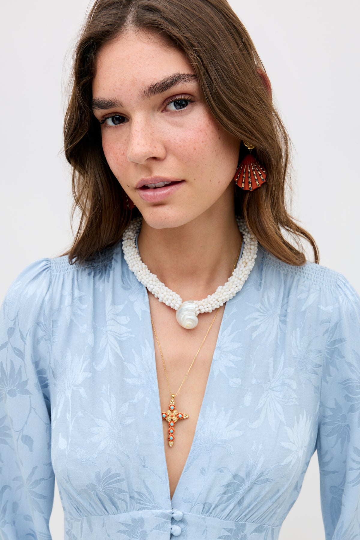 A woman wears the Lea - Daisy Jacquard Bluebell dress, paired with a white beaded necklace featuring a pearl, red fan-shaped earrings, and a gold necklace adorned with a jeweled cross pendant. She gazes at the camera with a neutral expression.