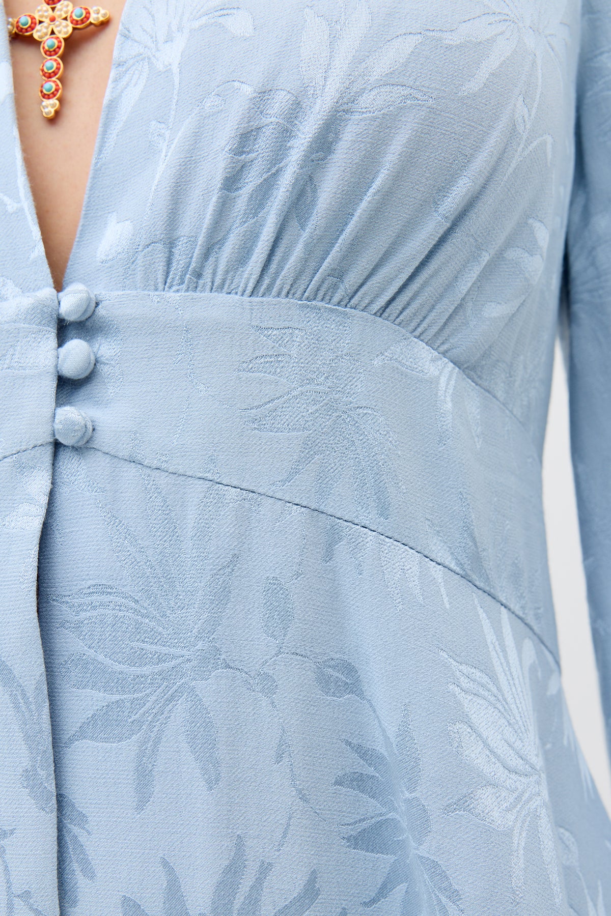 Close-up of the Lea - Daisy Jacquard Bluebell dress, featuring light blue jacquard fabric with subtle floral patterns and three fabric-covered buttons on the front; part of a gold and gemstone necklace is visible at the top.