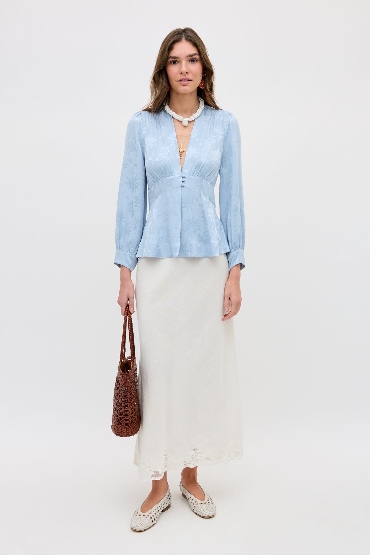 A woman stands against a white background wearing the Lea - Daisy Jacquard Bluebell blouse, paired with a long white skirt, white shoes, and holding a brown woven handbag. Her hair is loose and she has a neutral expression.