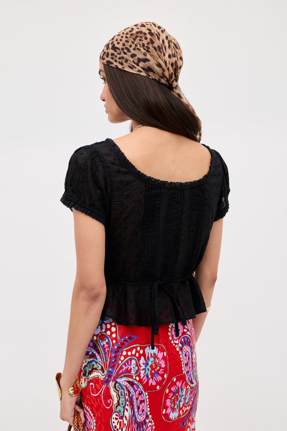 A woman is seen from behind wearing the Sandi - Black broderie anglaise cotton blouse, a red paisley skirt, a leopard print headscarf, and holding a small woven bag in her left hand—an effortless look for everyday style.