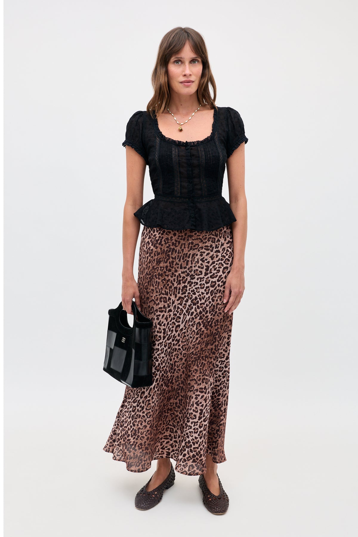 A woman is shown wearing a black short-sleeve blouse paired with the Kelly - Leopard Brown skirt, crafted from luxurious silk crepe de chine. She holds a black handbag, wears dark shoes and a necklace, and stands against a plain white background.