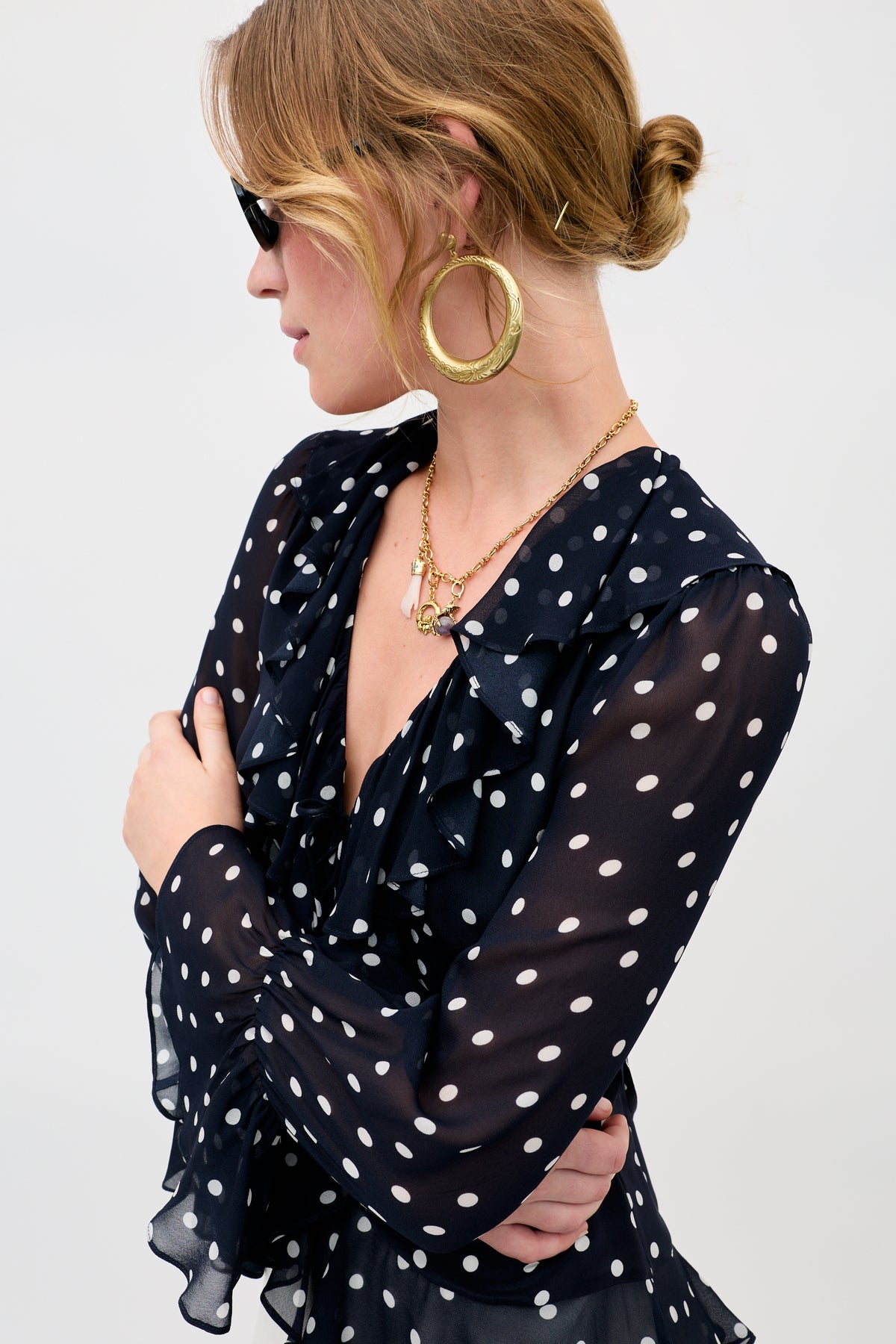 Woman with light brown hair in a low bun wears the Sian - Gold hoop earrings, layered gold necklaces, black sunglasses, and a navy sheer polka dot blouse, looking to the side with arms crossed.