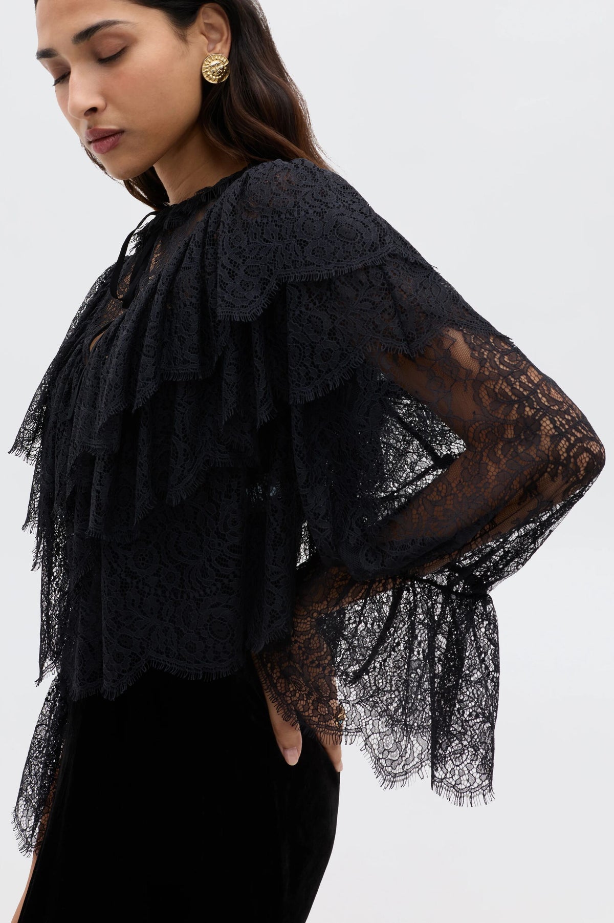 A woman with long dark hair and gold earrings stands sideways against a plain background, wearing the Faryn - Black, a sheer black lace ruffle blouse with long sleeves; her hand is on her hip.