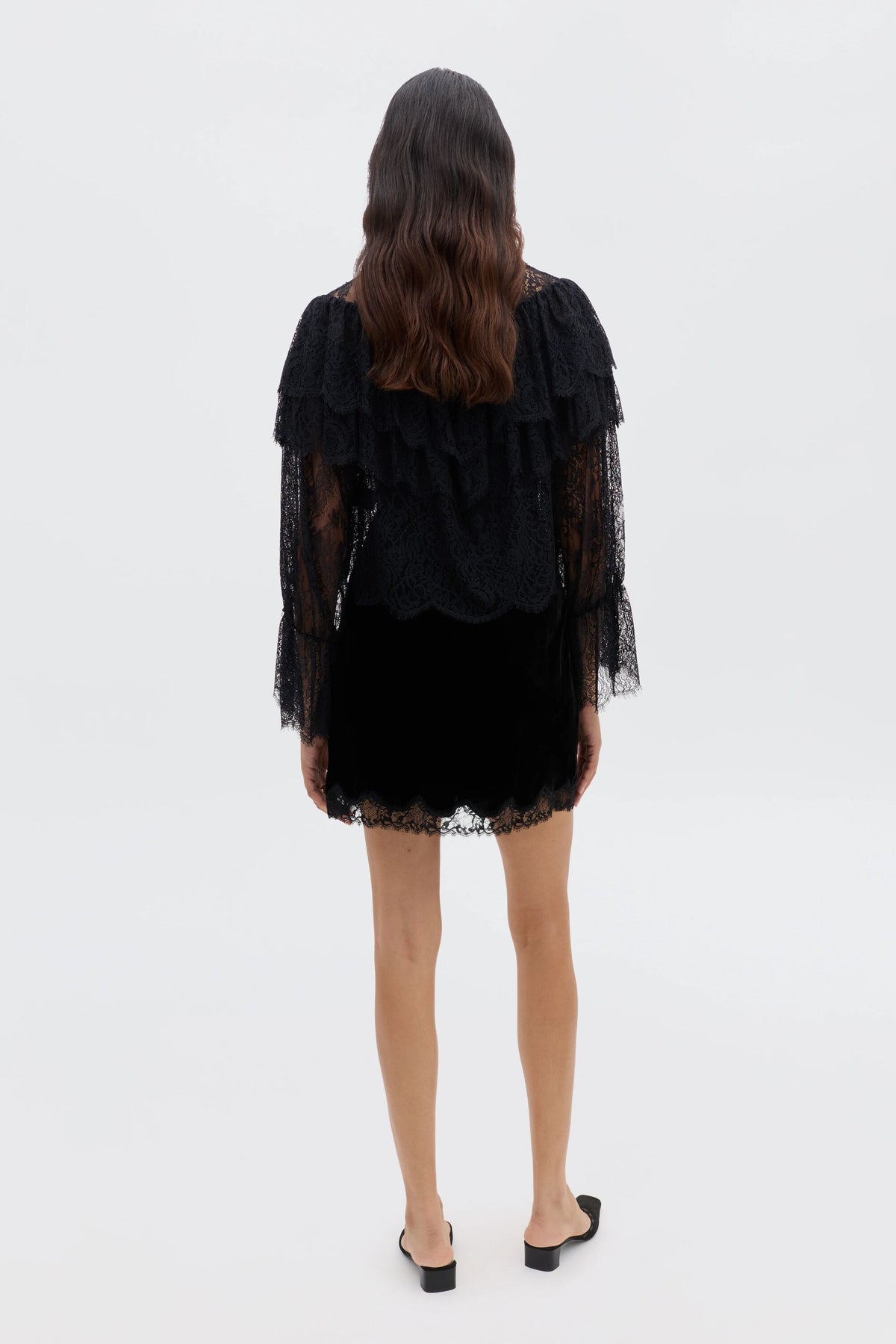 A woman with long wavy hair faces away, wearing a black lace ruffle blouse with tiered ruffles and long sleeves, paired with Faryn - Black open-back slip-on shoes, set against a plain white background.