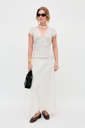 A woman stands against a white background wearing the Karia - Layla Dot Ivory blouse, paired with a long white skirt, brown woven shoes, black sunglasses, gold jewelry, a black bag, and a white beaded necklace.