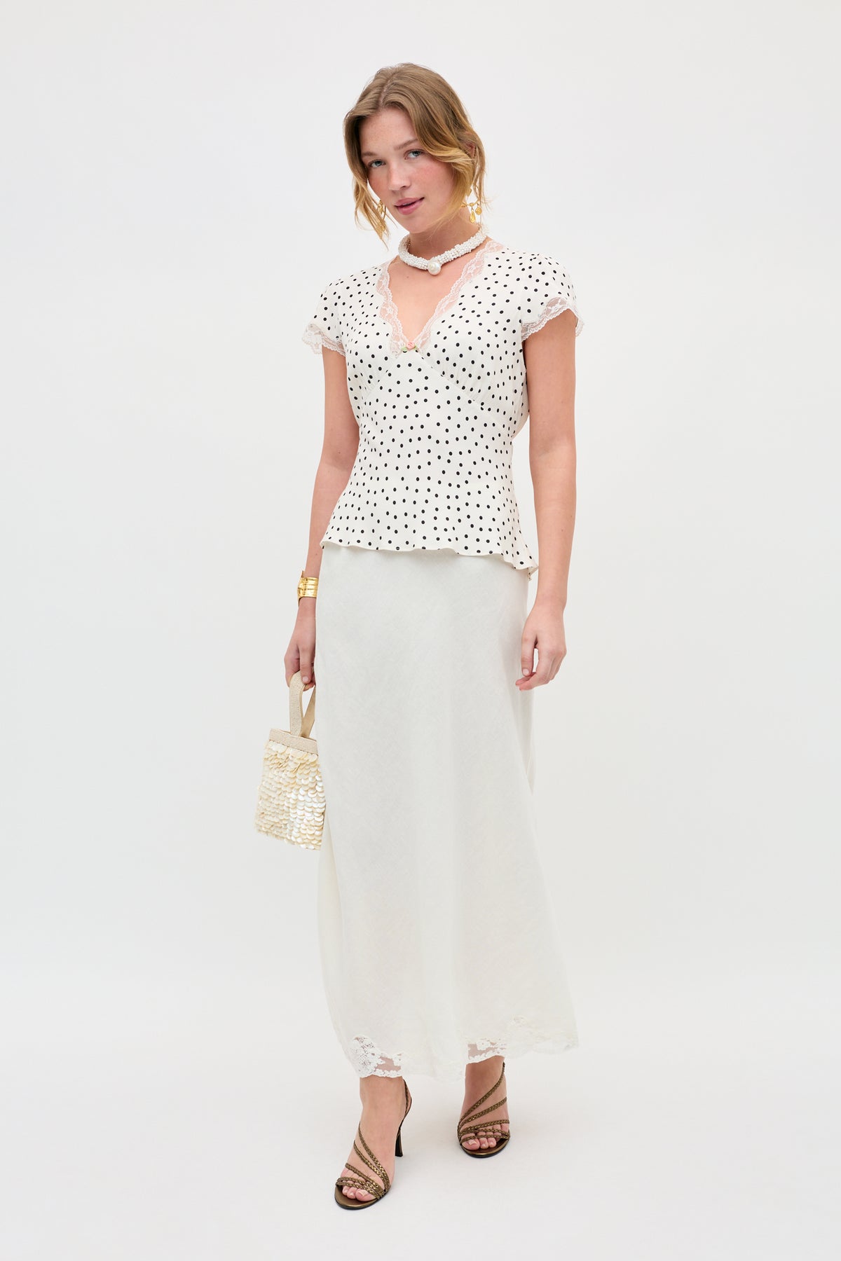 A woman stands against a white background wearing the Karia - Layla Dot Ivory blouse, a long white skirt, strappy heels, and holding a woven handbag, accessorized with a gold bracelet and a white beaded necklace.