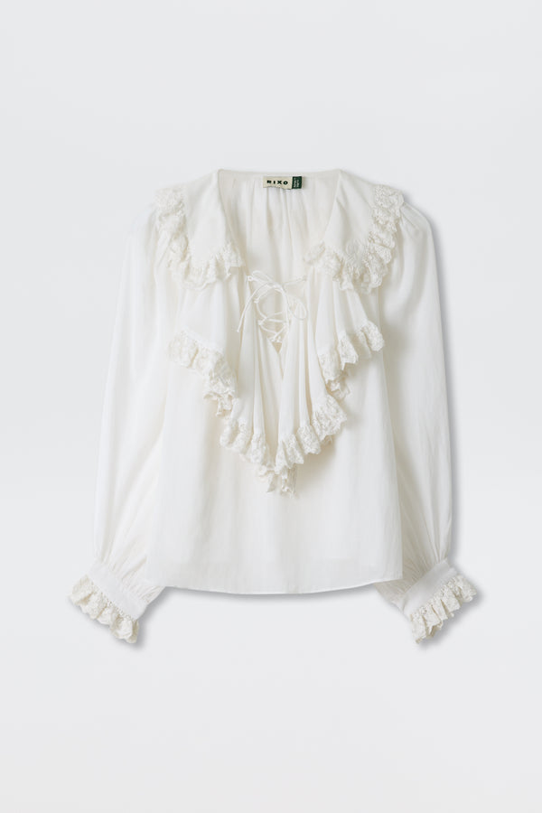 The Milia - White is a loose-fit, long-sleeved blouse featuring ruffled lace trim on the collar, chest, and cuffs, with a tie closure at the neckline.