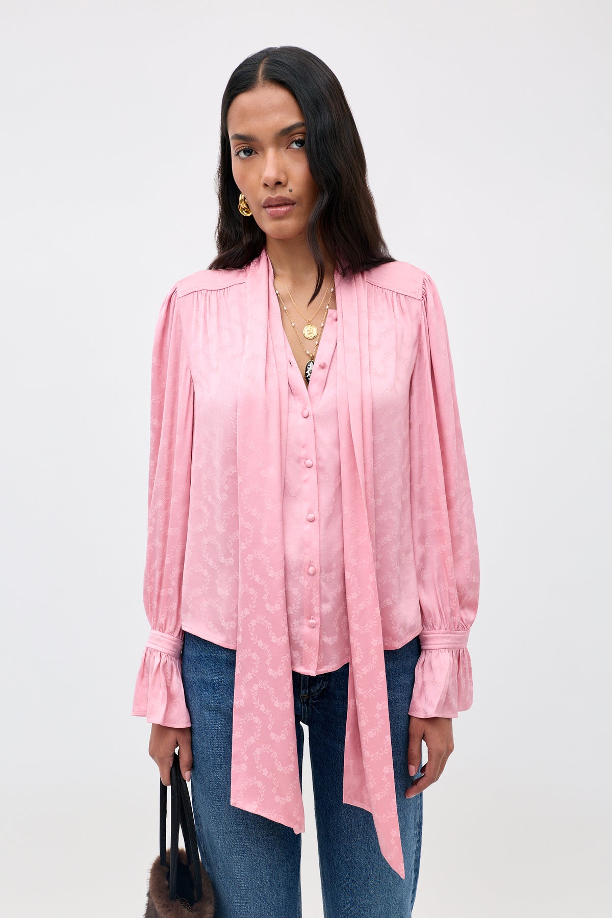 A woman wearing the Evanora - Floral Trail pink, long-sleeved blouse with a tie-neck detail and blue jeans stands against a plain light background, holding a handbag.