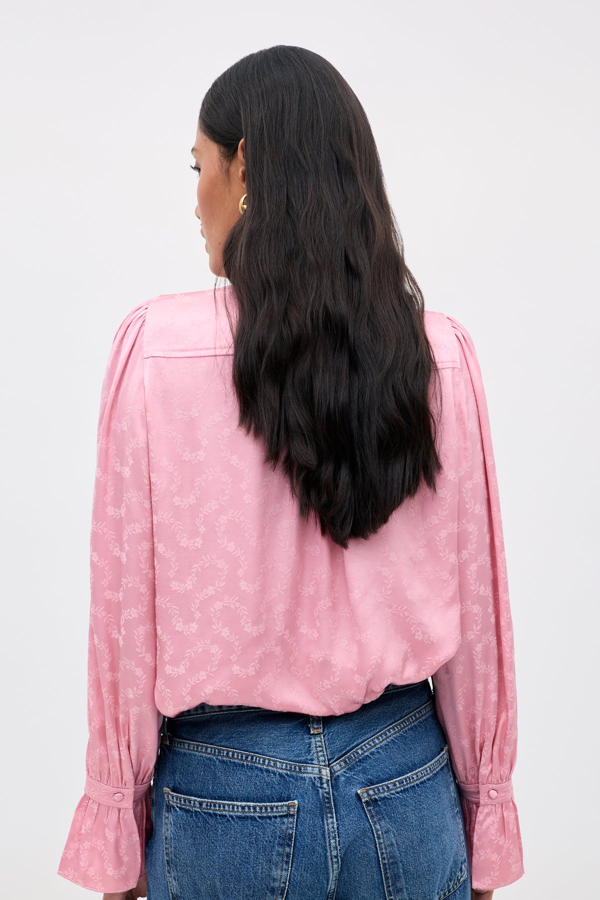 A person with long, wavy dark hair is seen from behind against a plain light background, wearing the Evanora - Floral Trail long-sleeve blouse in light pink paired with blue jeans.