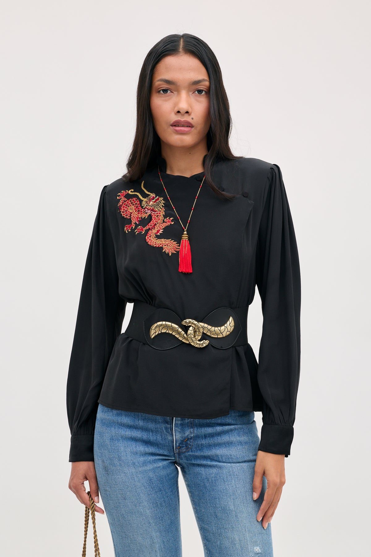 A model wears a Sylvie Rose - Dragon Embroidery Red blouse with red tassel necklace and wide black belt with gold clasp, paired with blue jeans.