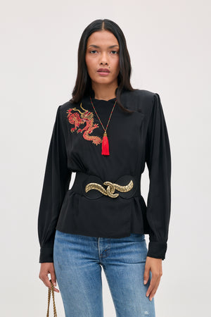 A model wears a Sylvie Rose - Dragon Embroidery Red blouse with red tassel necklace and wide black belt with gold clasp, paired with blue jeans.