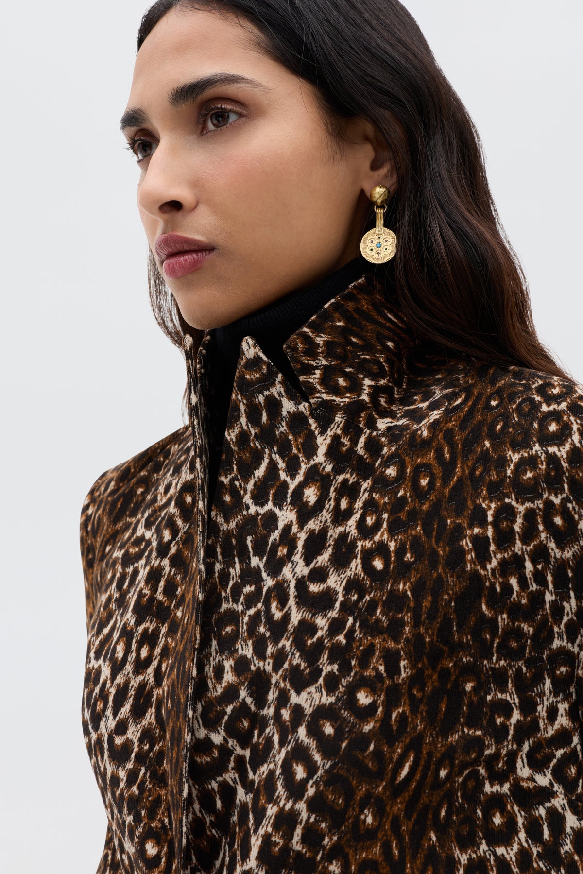A woman with long dark hair wears the vintage-inspired Xander - Lyra Leopard Black jacket, paired with a black turtleneck and gold dangling earrings, standing against a plain light gray background.