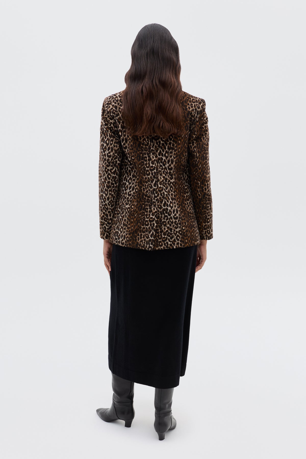 A person with long wavy hair stands facing away, wearing the Xander - Lyra Leopard Black jacket, a long black skirt, and knee-high black boots against a plain white background.