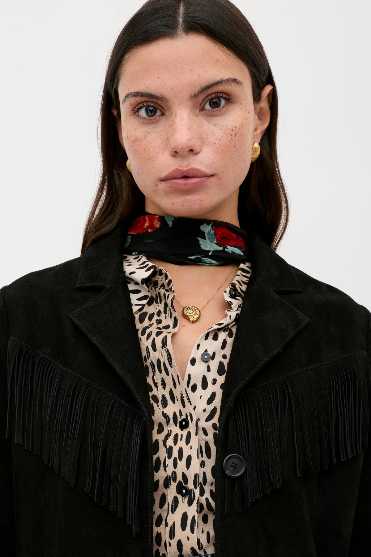 A woman with long brown hair and freckles gazes at the camera, wearing gold jewelry, a red floral black scarf, and the Teagan - Black jacket over a spotted blouse.
