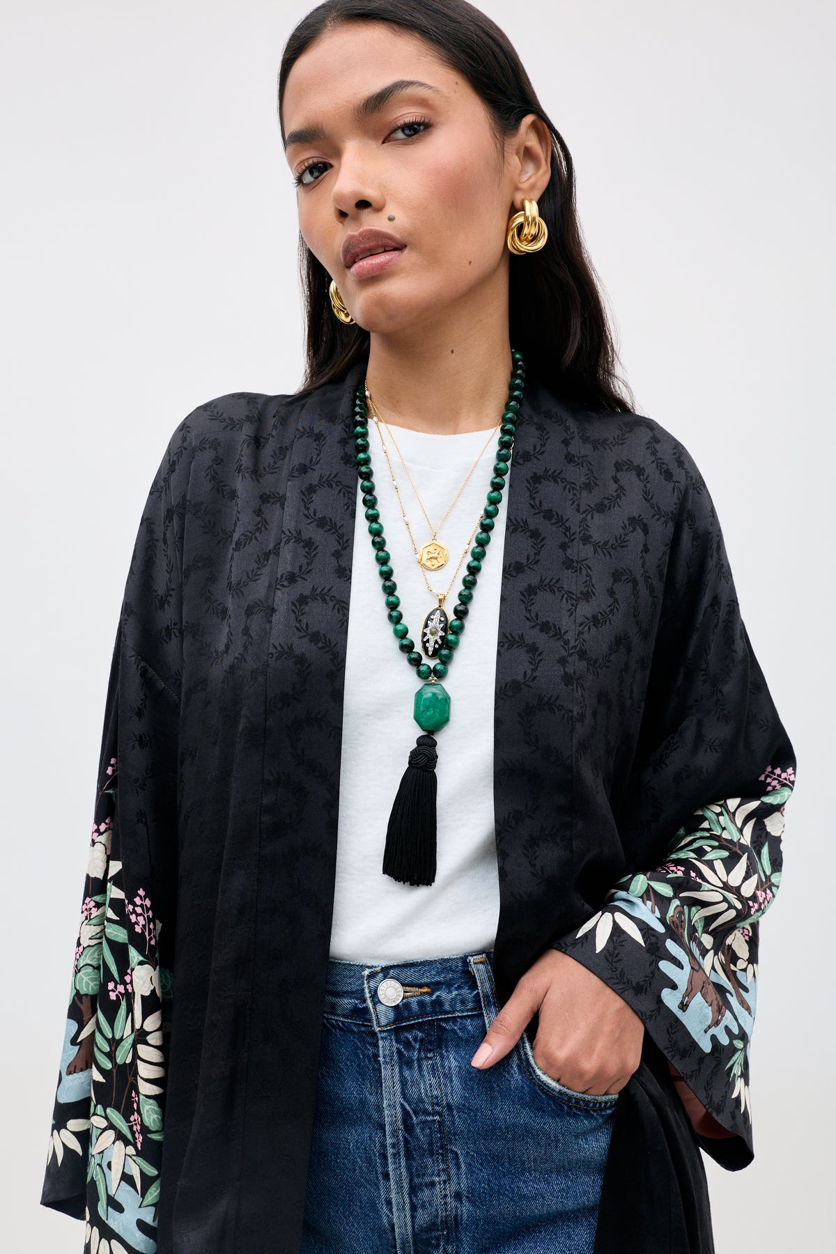 Wearing the Kaiden - Blossom Jungle Black, a patterned black kimono with floral sleeves, gold earrings, layered necklaces, and a beaded tassel necklace, a woman stands confidently with one hand in her jeans pocket and gazes directly at the camera against a plain background.