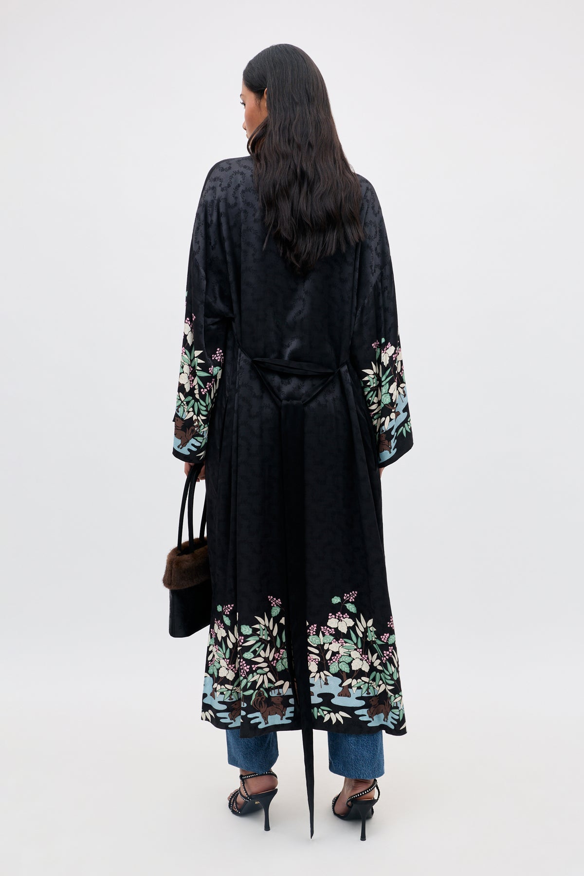 A woman with long dark hair, seen from behind, wears the Kaiden - Blossom Jungle Black floral robe over jeans and heels, holding a black handbag.