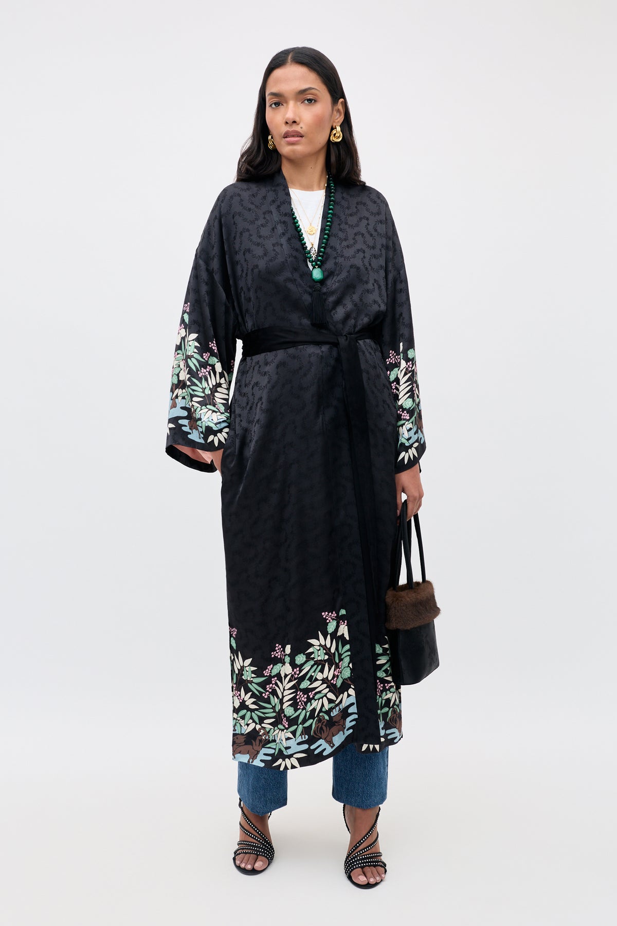 A woman wearing the Kaiden – Blossom Jungle Black long kimono with floral patterns at the hem and sleeves, paired with blue jeans, strappy sandals, layered necklaces, and carrying a black handbag stands against a plain white background.