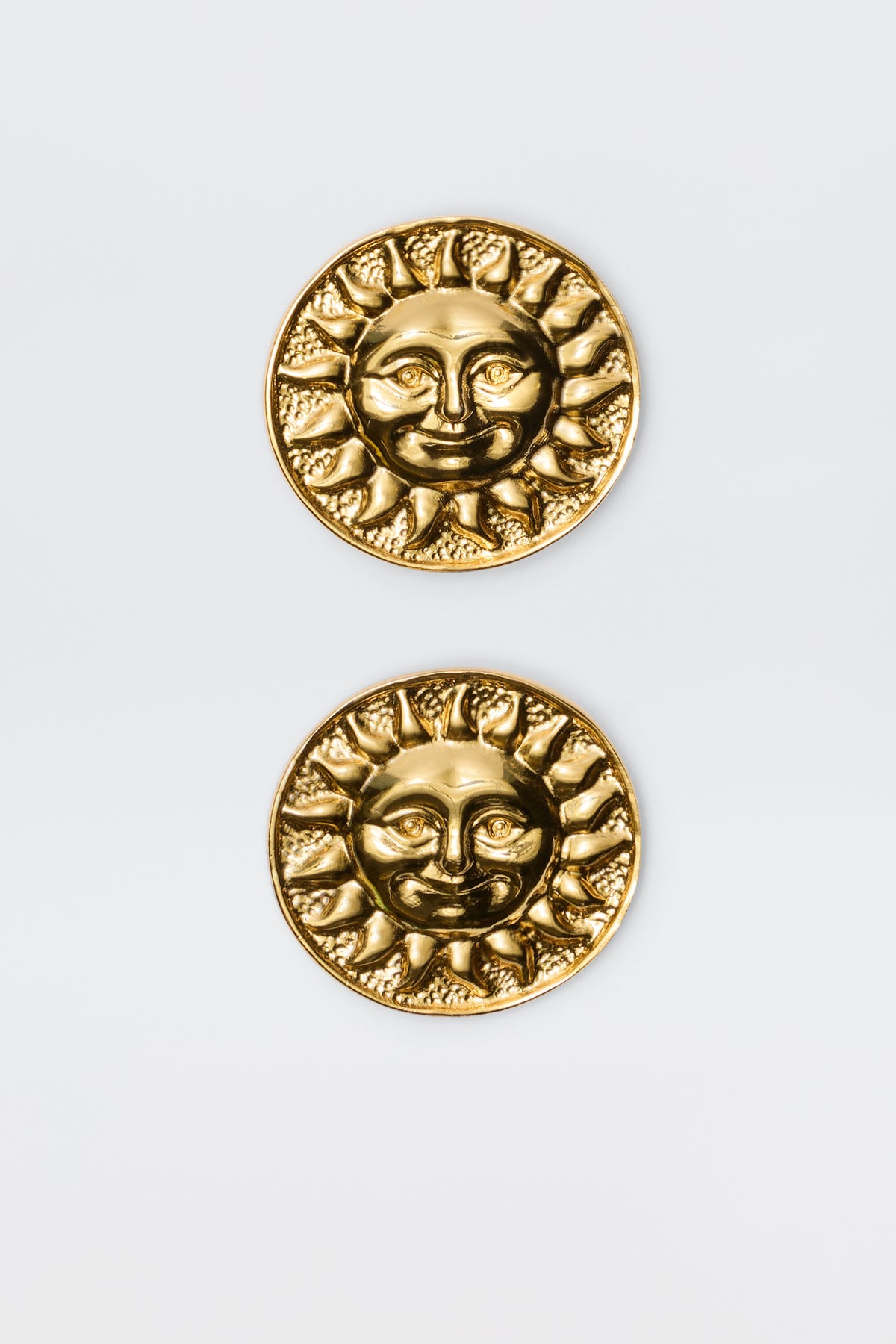 Introducing Noni - Gold: round gold-plated brass coins with embossed sun faces, rays, and intricate details. These vintage-inspired pieces are displayed vertically on a light gray background, resembling elegant round stud earrings.