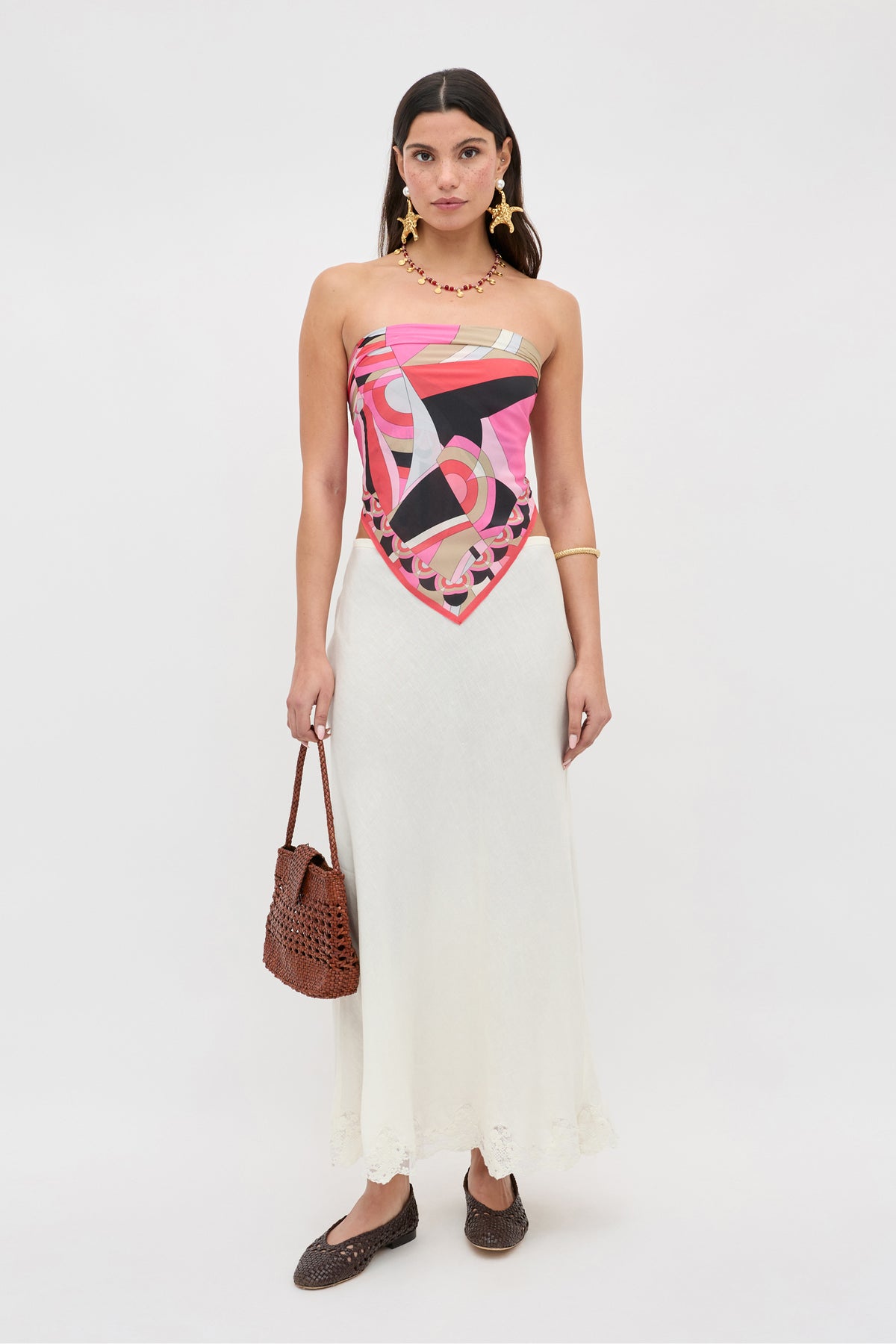 A woman stands against a white background wearing the Menno - Soho Geo Pink, a colorful pink patterned top. She pairs it with a long white skirt, brown woven shoes, gold jewelry, and carries a brown woven handbag. Her look is completed with a vibrant silk scarf elegantly draped around her neck, making for the perfect fashion accessory.
