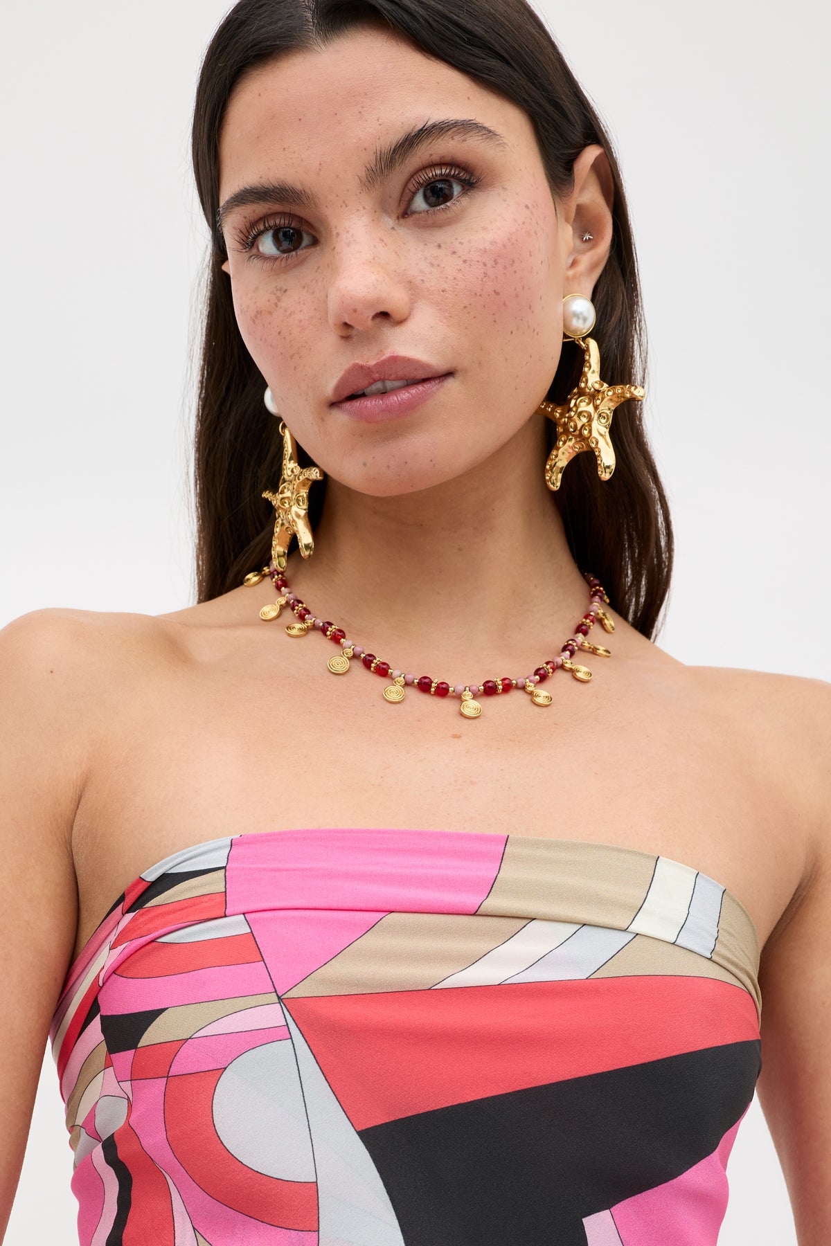A woman with long dark hair wears the Menno - Soho Geo Pink, a colorful strapless top, paired with gold star-shaped earrings adorned with pearls and a red beaded necklace featuring gold discs. She elegantly styles her look with a silk scarf as a chic accessory, standing against a plain light background.