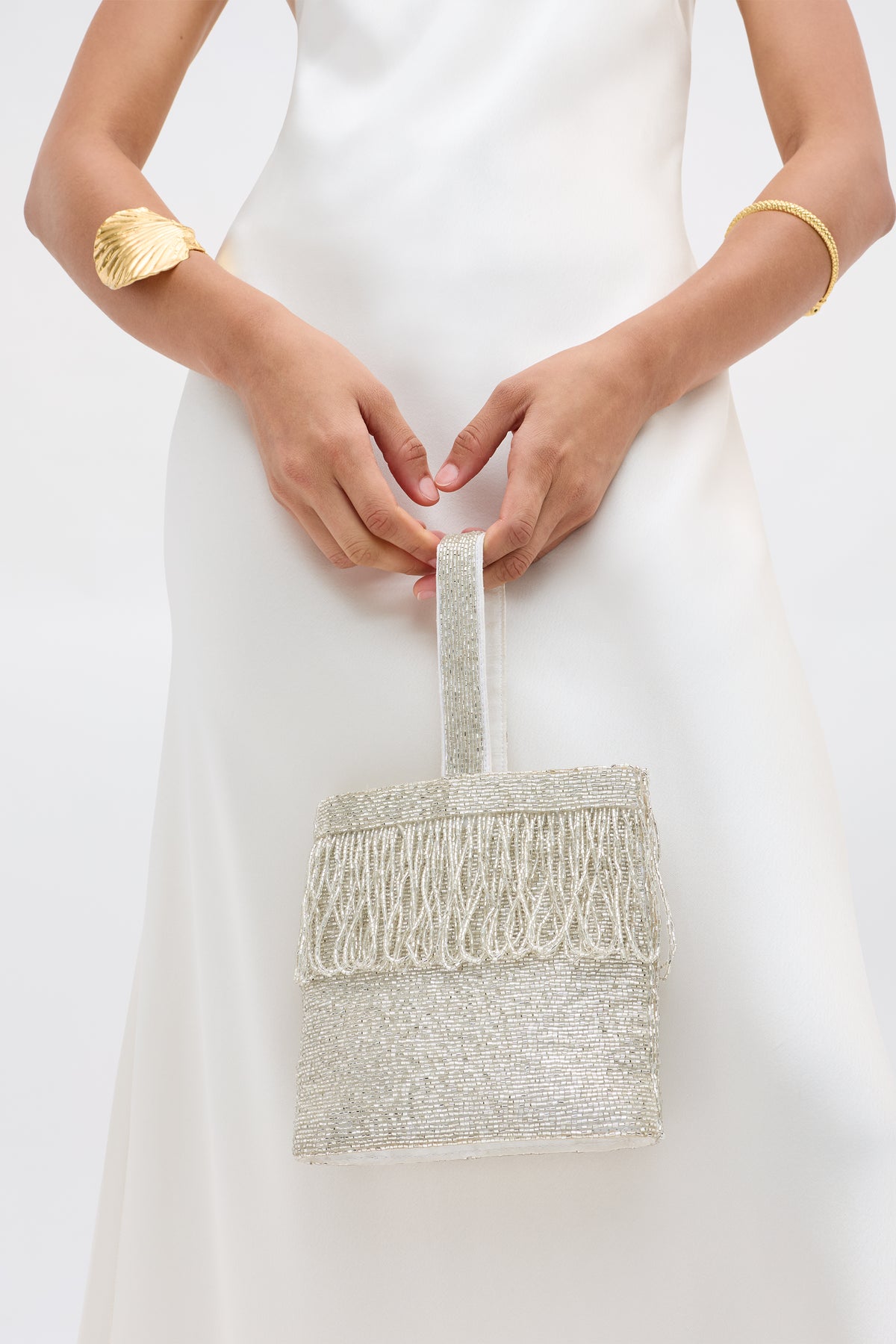 Wearing a white satin dress, a person holds the Zenni - Beaded Silver bag with fringe and a rounded top handle, styled with a gold shell bracelet and simple gold bangle, set against a plain light background.