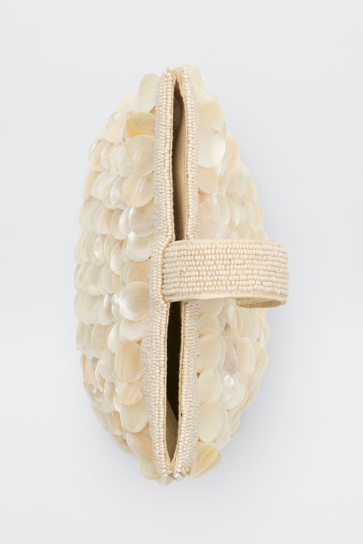 The Zenni - Shell clutch is beige with shiny, overlapping shell-like discs and a beaded handle across the opening, shown from the side against a plain white background.