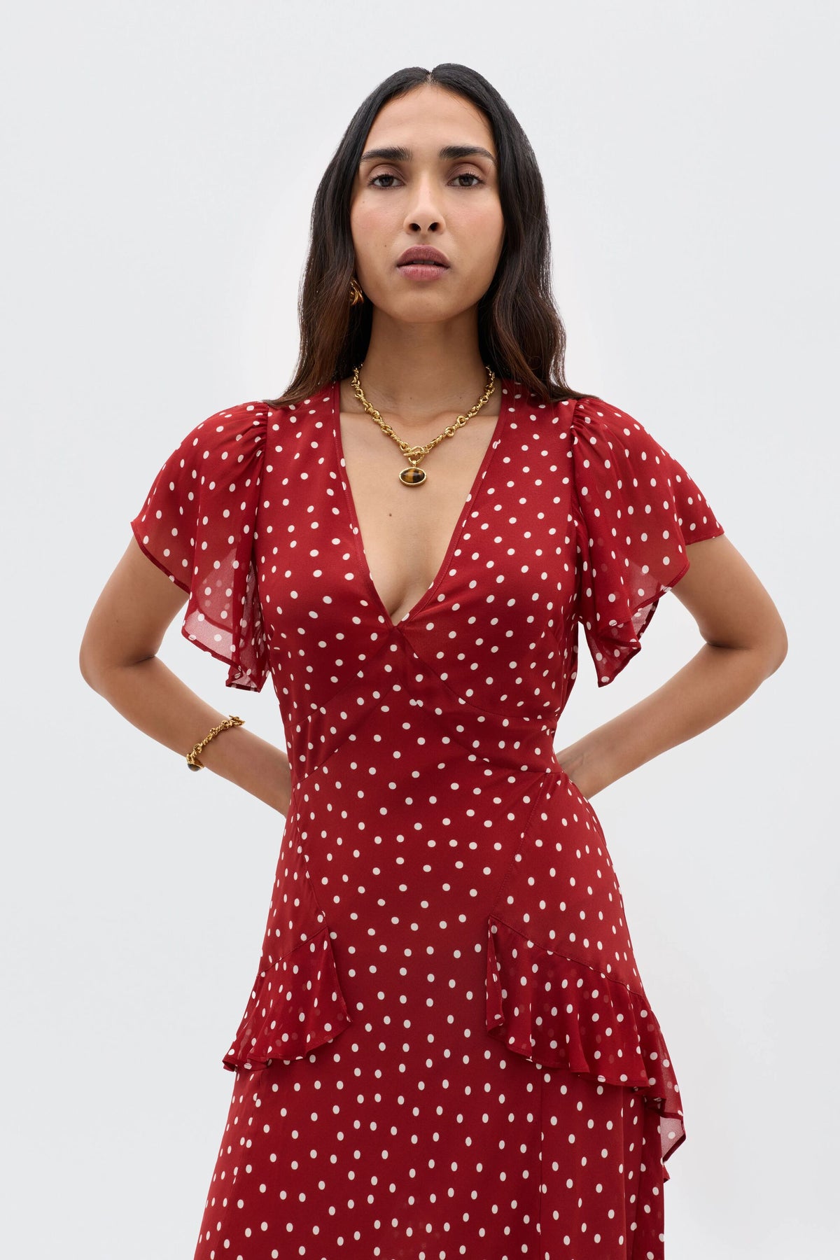 A woman with long dark hair wears the Evie - Layla Dot Red dress with short flutter sleeves and a V-neckline. She accessorizes with gold hoop earrings, a necklace, and a bracelet, standing against a plain light background.