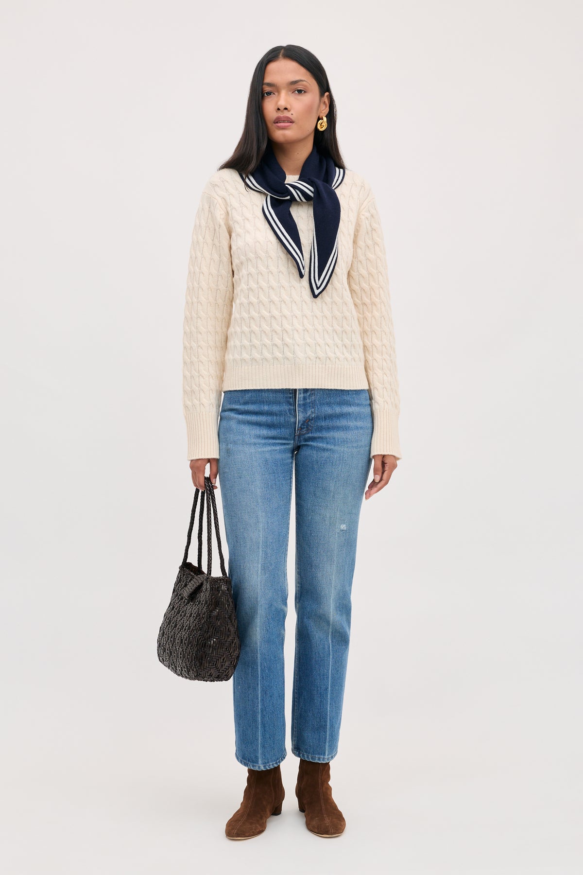 A person wears a cream cable-knit sweater with a Lianora - Sailor Stripe Navy neck scarf, blue jeans, a dark woven handbag, and brown suede boots.
