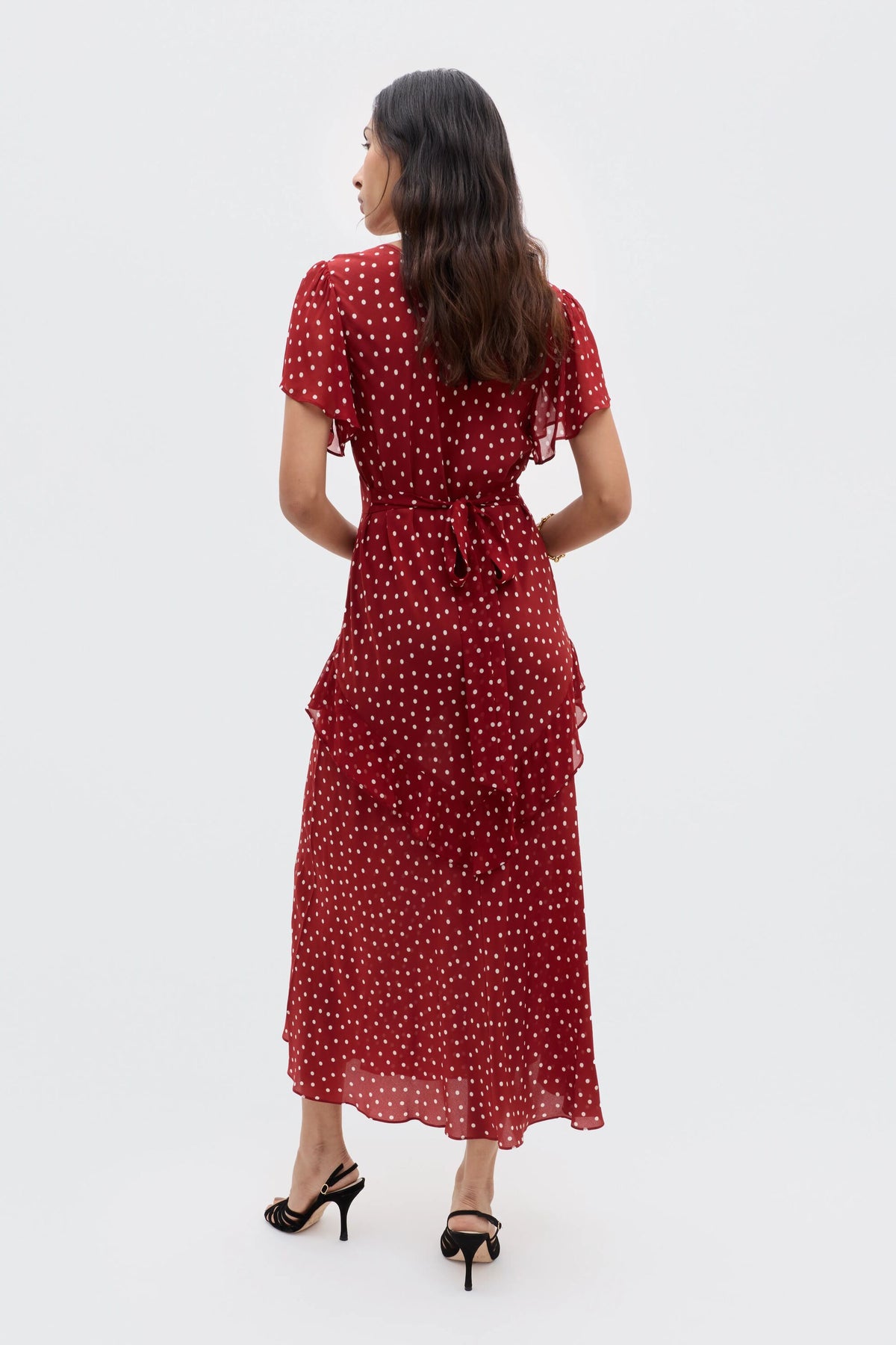 A woman stands with her back to the camera, wearing the RIXO Evie - Layla Dot Red dress featuring white polka dots and ruffled layers, elegantly paired with black strappy heeled sandals.