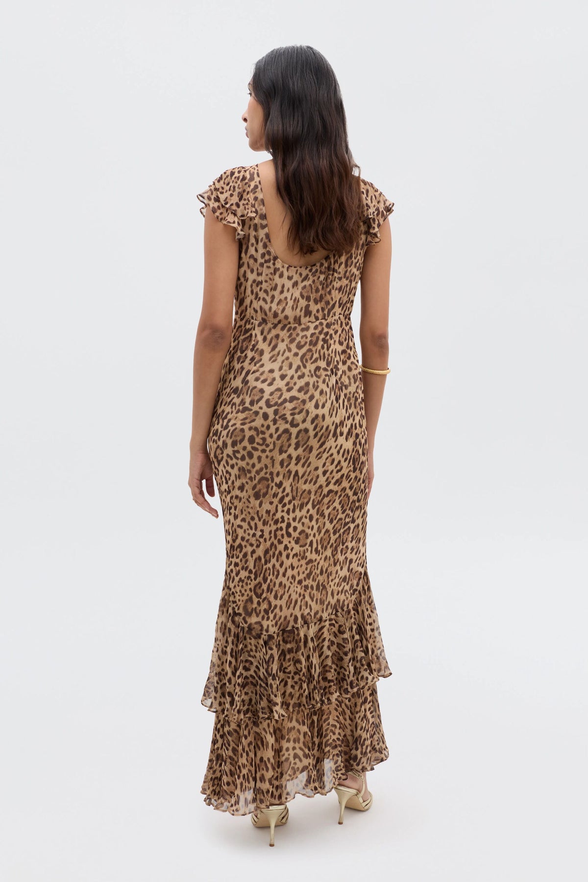 A woman with long dark hair models the Ordinia - Mila Leopard Brown gown, a sleeveless, ankle-length dress with a ruffled hem and heels, standing with her back to the camera against a plain white background—ideal for black-tie weddings.