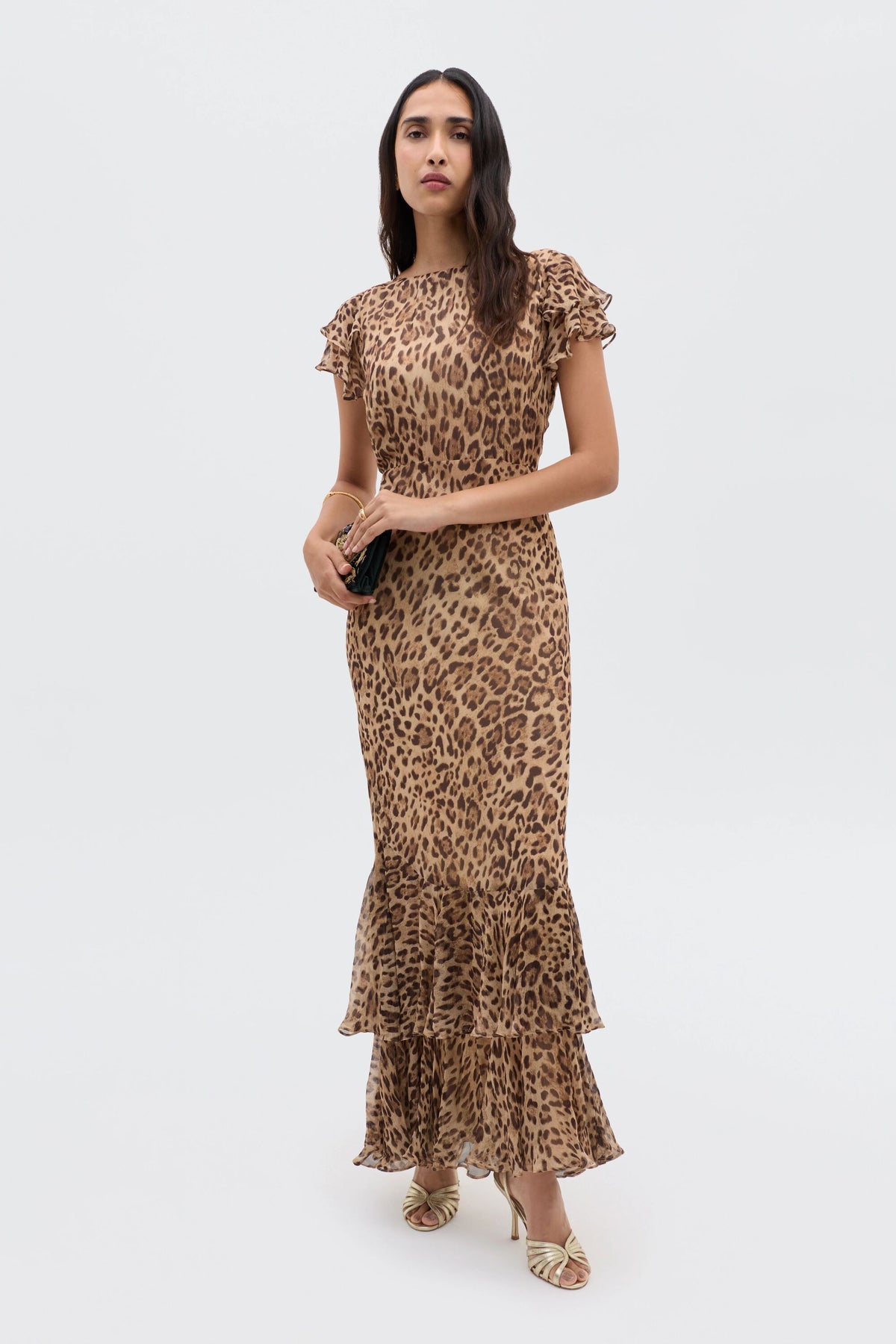 A woman stands against a plain background wearing the fitted, ankle-length Ordinia - Mila Leopard Brown gown with ruffled sleeves and hem, styled with gold sandals and a small black clutch—ideal for black-tie weddings.