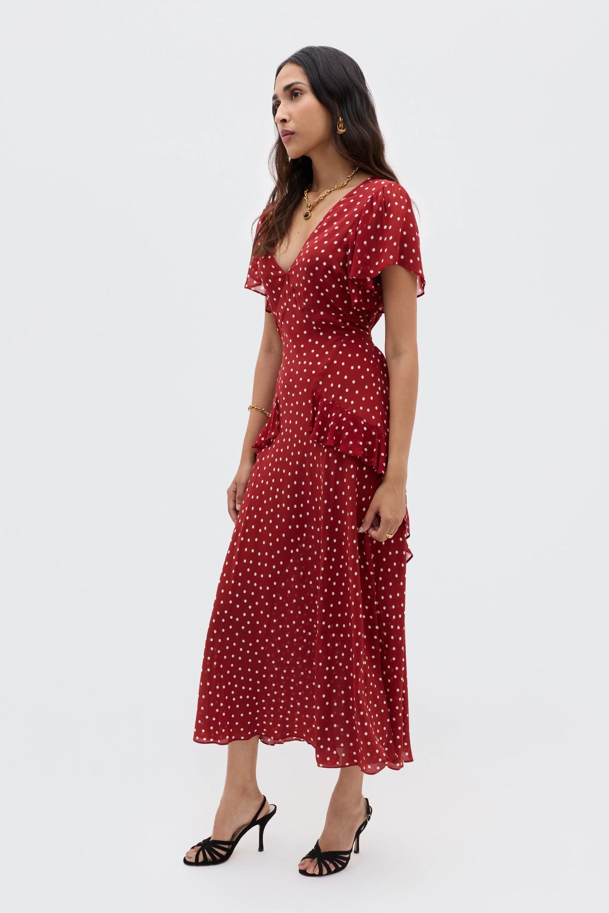A woman in the RIXO Evie - Layla Dot Red dress with short sleeves, v-neck, and ruffle details stands in heeled sandals against a plain white background.