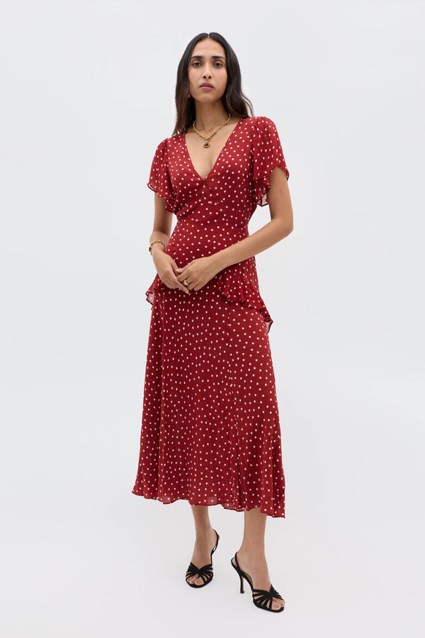 A woman wears the Evie - Layla Dot Red dress—red, short-sleeved, V-neck with polka dots—paired with black heeled sandals, standing against a plain light gray background.