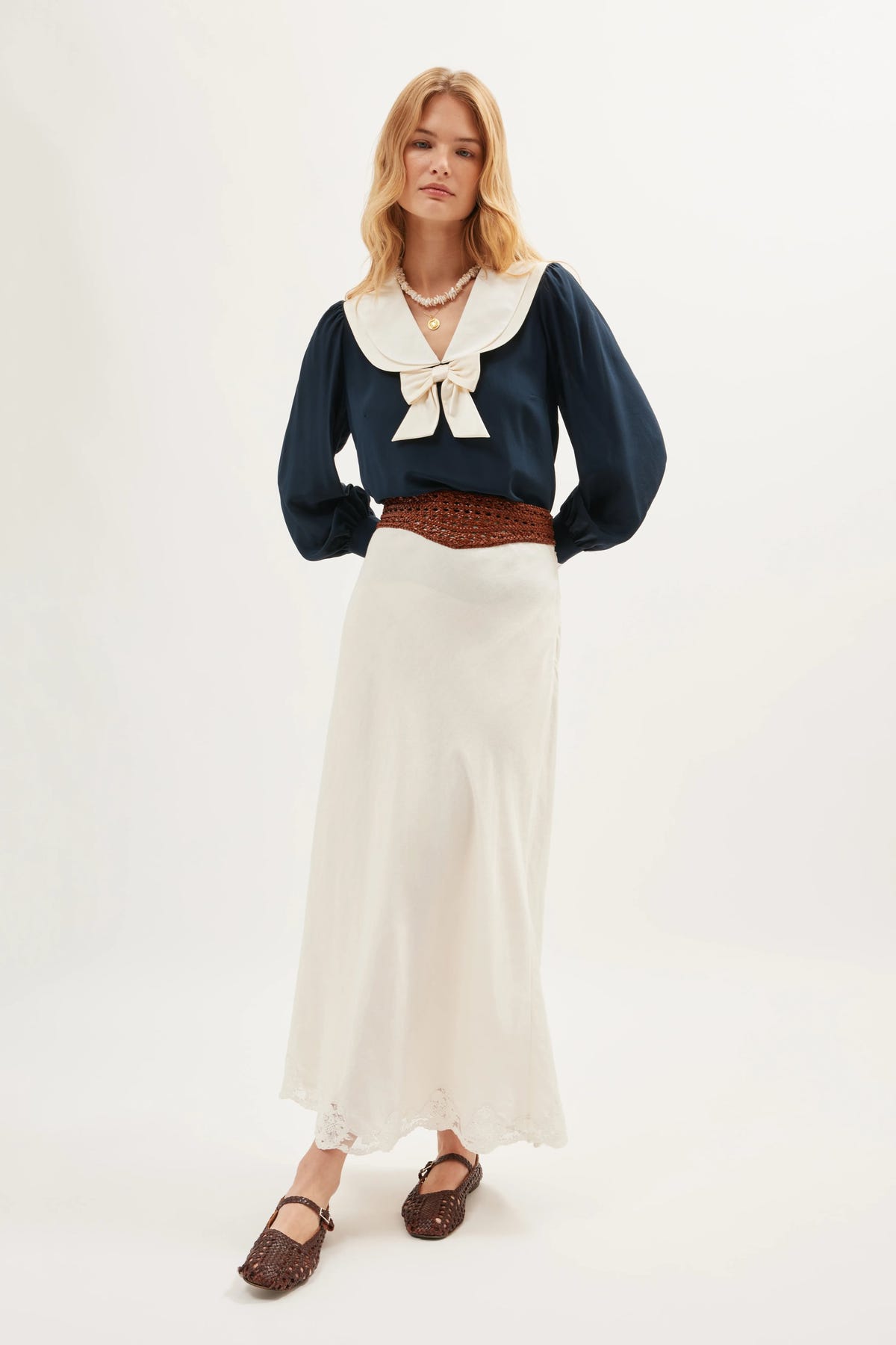 A person wears the Crystal - Cream, a long ivory linen skirt, paired with a navy long-sleeve blouse with a large white collar and bow, brown woven belt and shoes, and layered necklaces, standing against a plain white background.