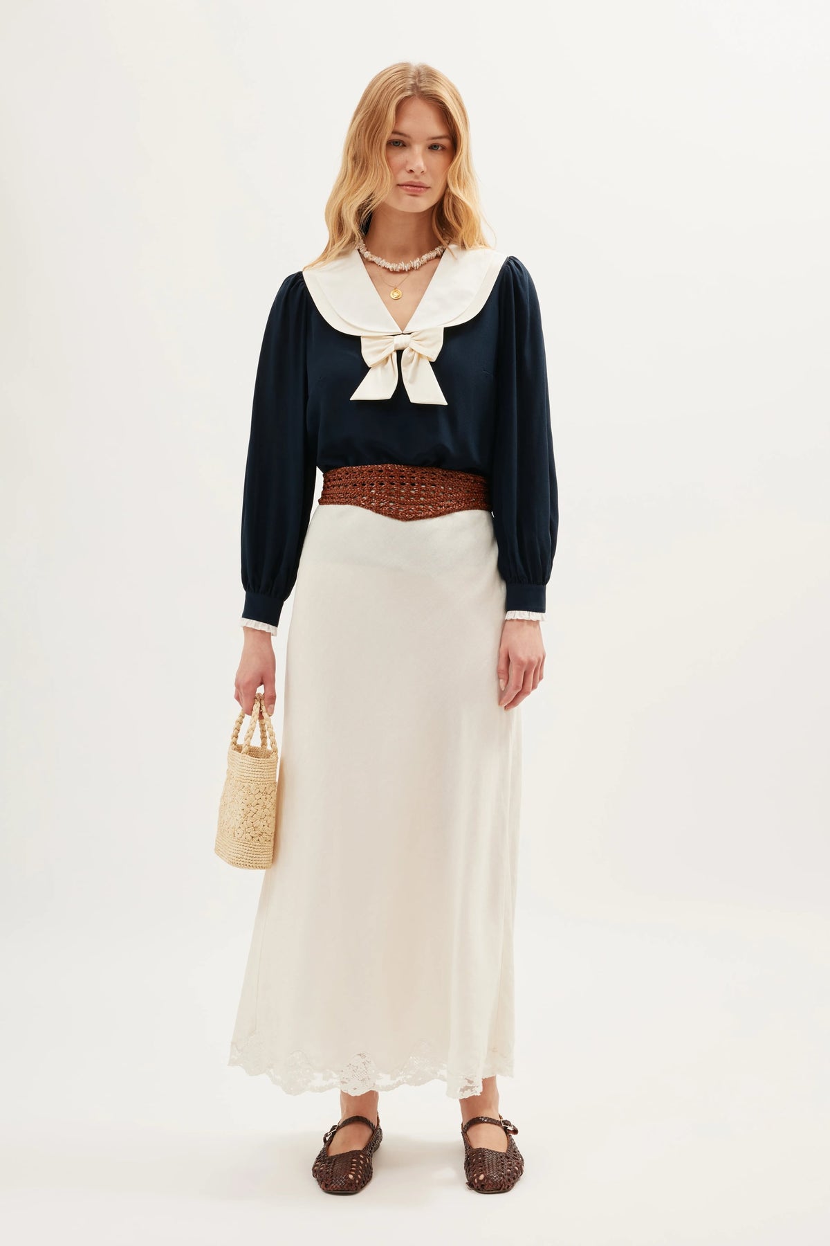 A woman stands against a white background wearing the Crystal - Cream linen skirt, paired with a navy blouse with a large white collar and bow, brown woven belt and shoes, and holding a woven handbag.