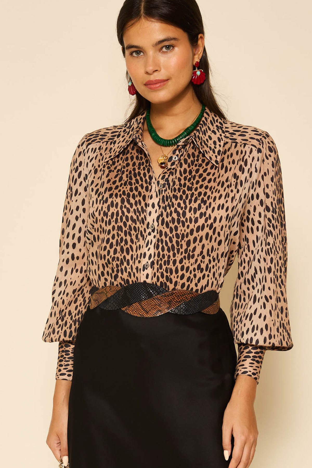 A woman poses against a neutral background wearing the Blake - Antelope Spot long-sleeved, button-up shirt tucked into a black skirt, styled with a green necklace and red earrings.