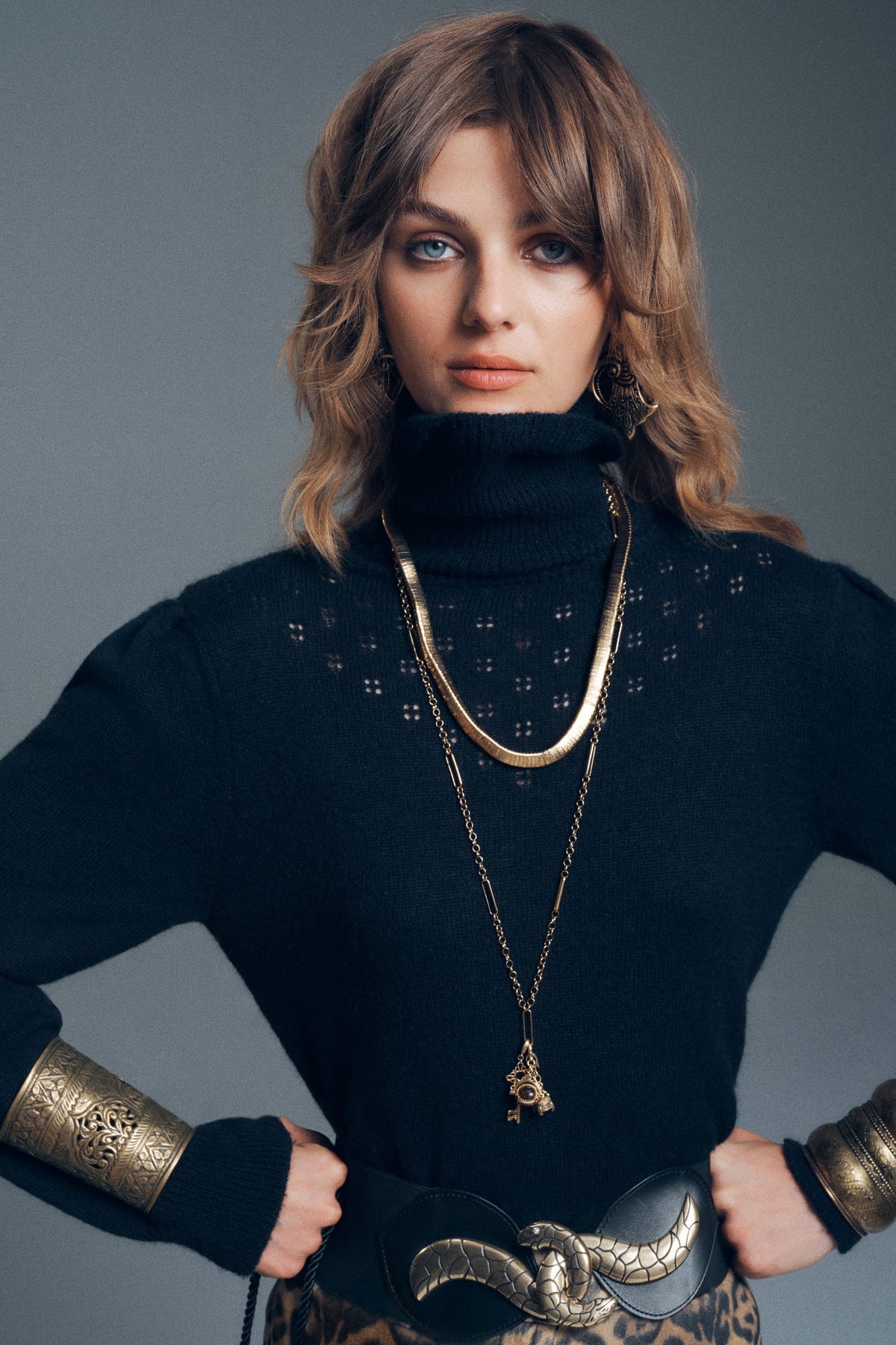 A person wearing a black turtleneck sweater and Lucian - Gold necklaces, including a thick flat chain and a long pendant chain, with an ornate gold cuff on their wrist and a snake-motif belt buckle.