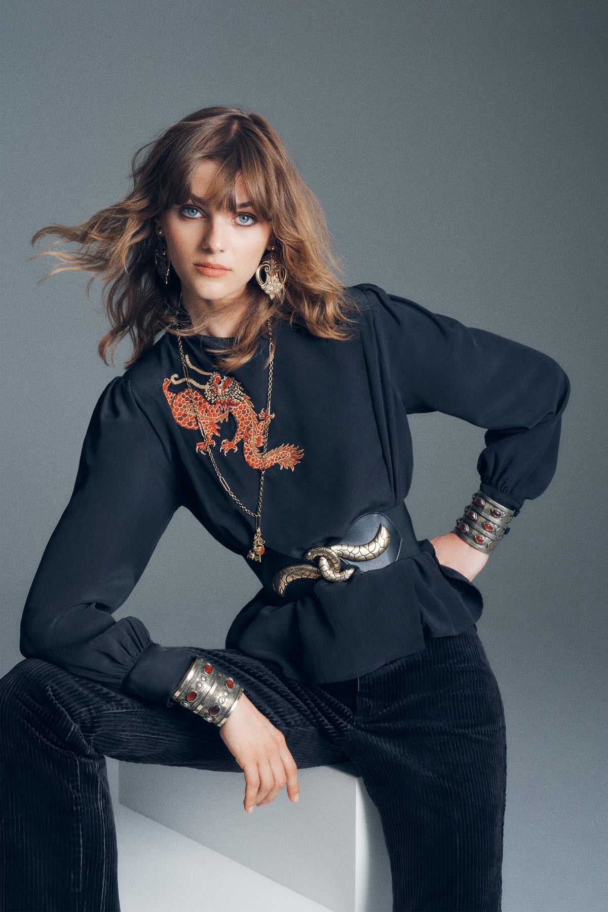 A person wearing a Sylvie Rose - Dragon Embroidery Red long-sleeved top with a silver snake-shaped belt and stacked metal cuffs with red stones.
