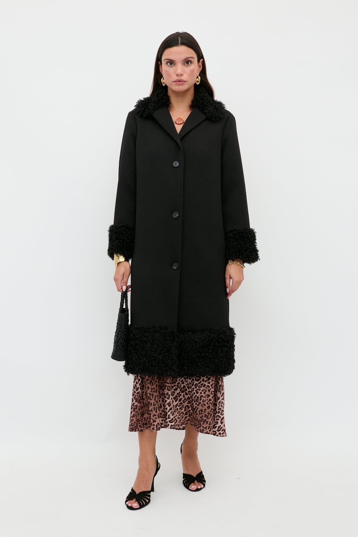A woman stands against a white background wearing the Diara - Black, a long black coat with textured faux-fur shearling trim at the cuffs and hem. She pairs it with a leopard print skirt, black heels, and carries a black bag—a timeless wardrobe staple for any season.