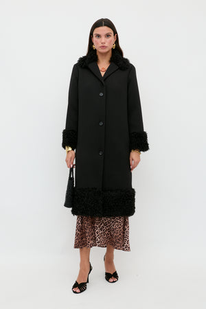 A woman stands against a white background wearing the Diara - Black, a long black coat with textured faux-fur shearling trim at the cuffs and hem. She pairs it with a leopard print skirt, black heels, and carries a black bag—a timeless wardrobe staple for any season.