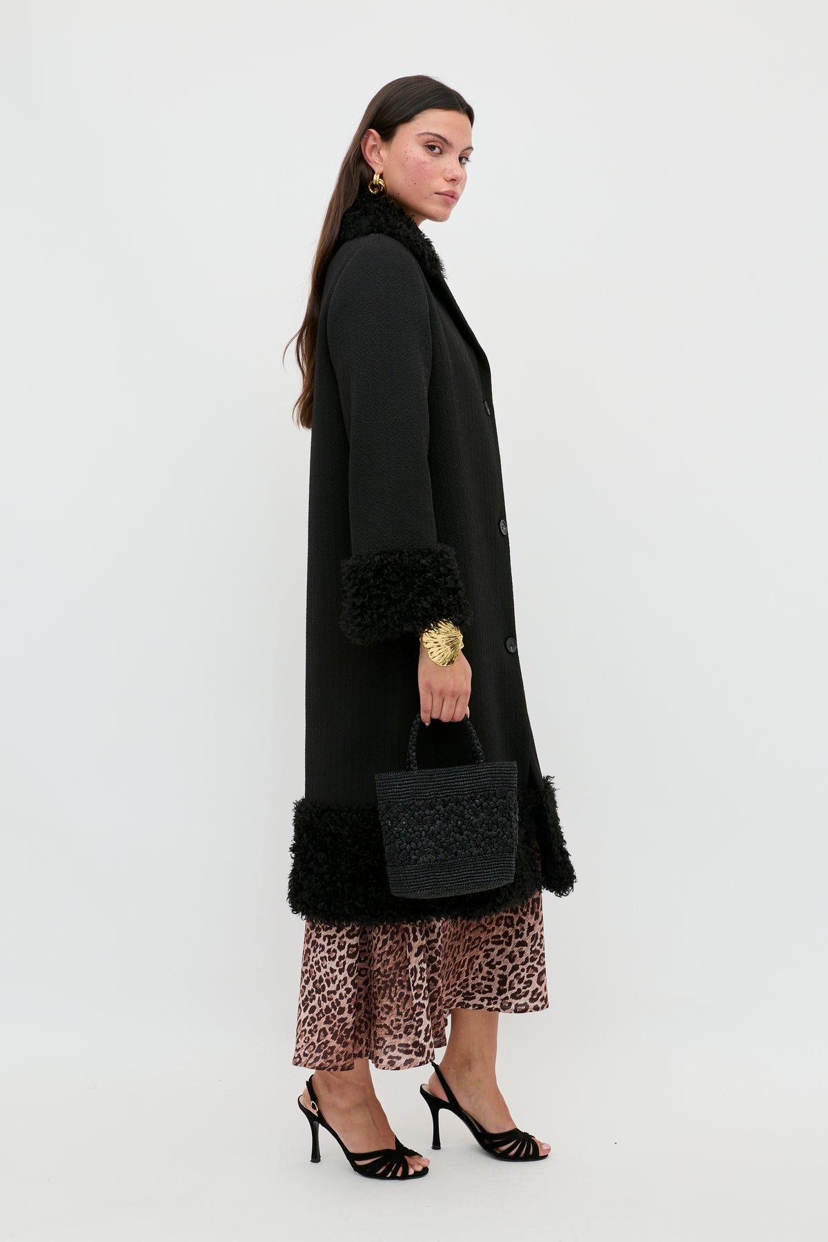 A woman wears the Diara - Black coat, a long black piece detailed with luxurious faux-fur shearling trim at the cuffs and hem. She carries a textured black handbag, pairs the look with a leopard print skirt and black high-heeled sandals, creating a striking wardrobe staple set against a plain white background.