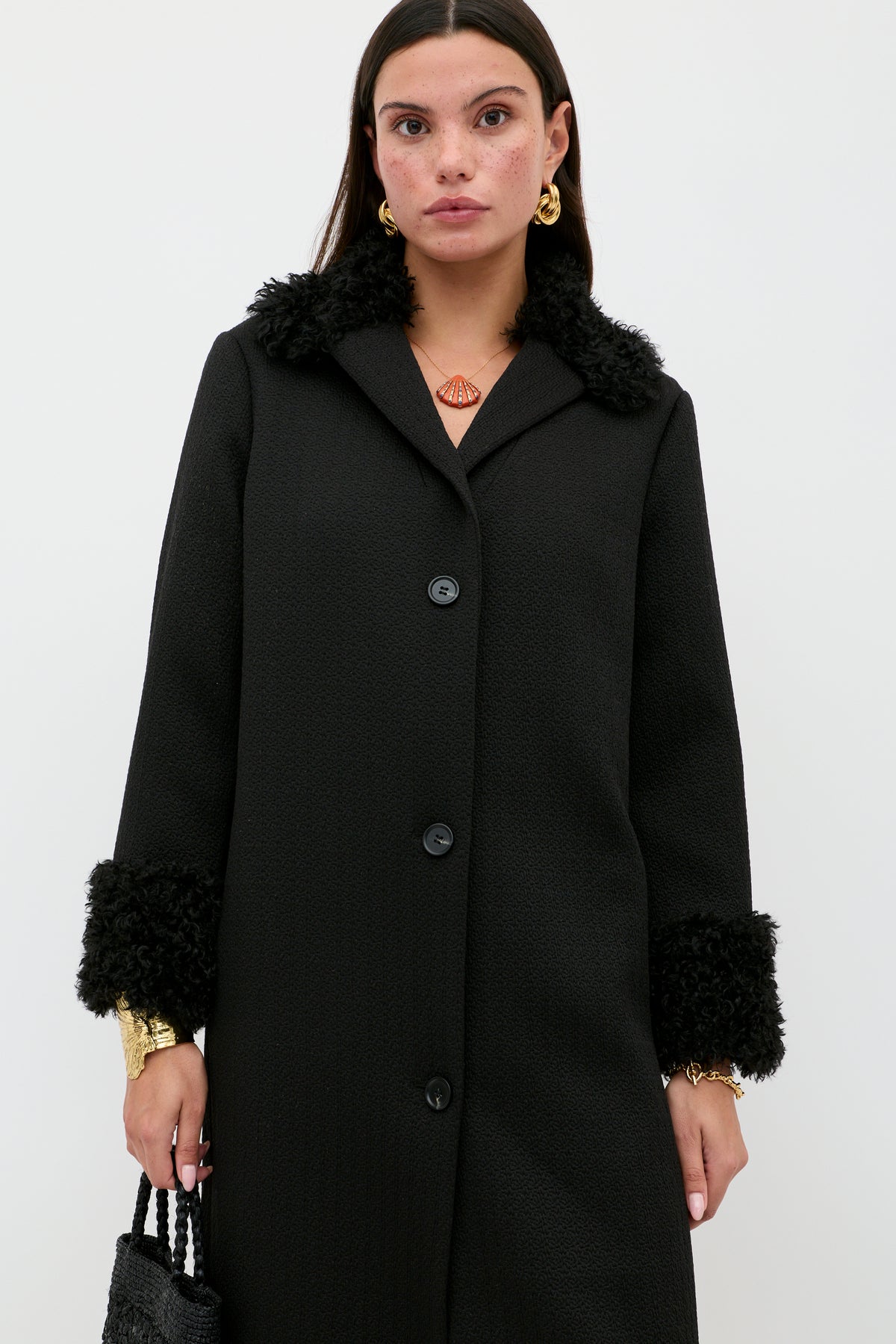 Wearing the Diara - Black, a long textured coat with faux-fur shearling trim on the cuffs and collar, gold jewelry, and holding a black bag, a woman stands against a plain white background—a true wardrobe staple for effortless elegance.
