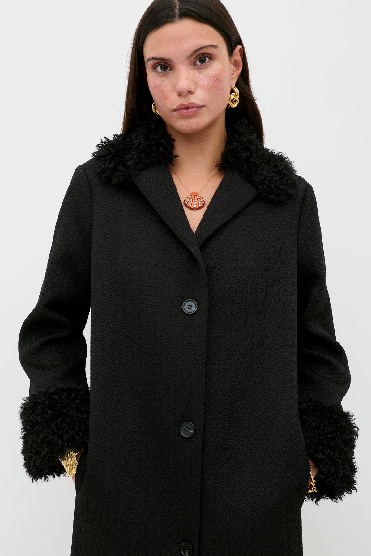A woman wearing the Diara - Black coat, a wardrobe staple with a textured black design and luxurious faux-fur shearling trim at the collar and cuffs, along with gold earrings and a geometric pendant necklace, stands against a plain white background.